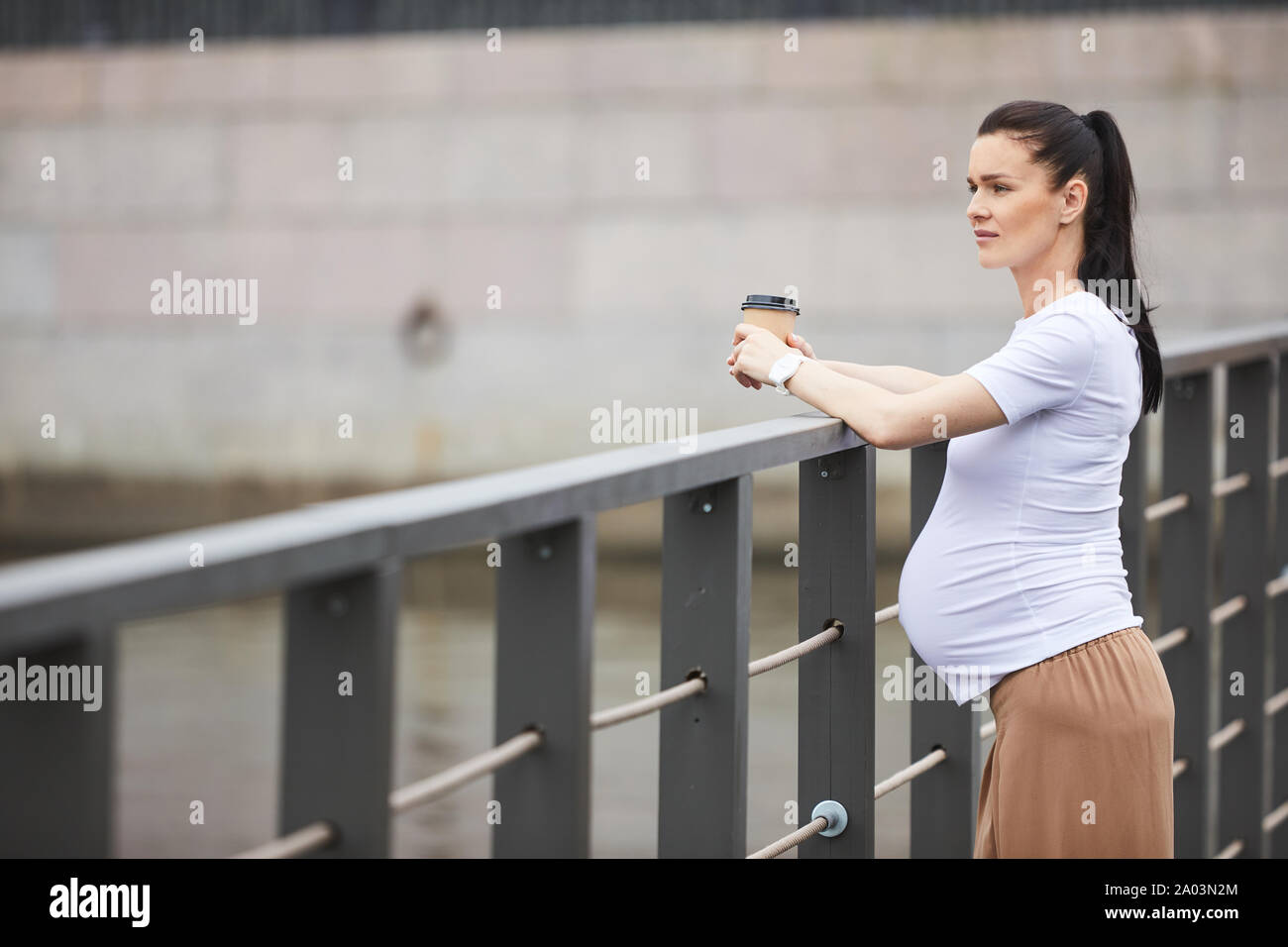 Jeune femme enceinte dans les vêtements élégants de boire du café et à l'écart avec pensive tout en se tenant debout sur le pont Banque D'Images