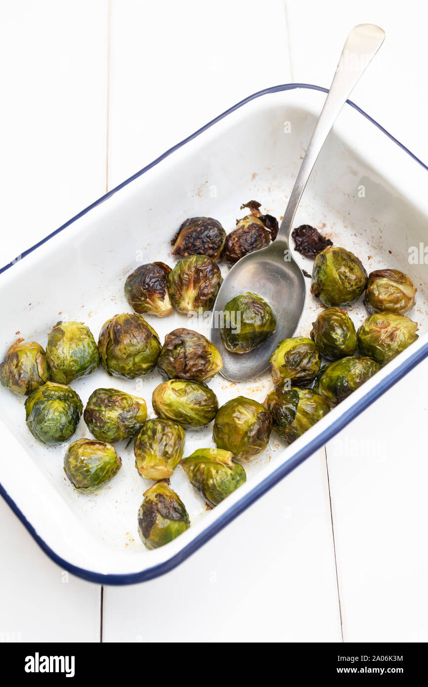 Brassica oleracea. Choux de Bruxelles rôtis dans un plat de cuisson en émail blanc avec une cuillère sur fond de bois blanc Banque D'Images