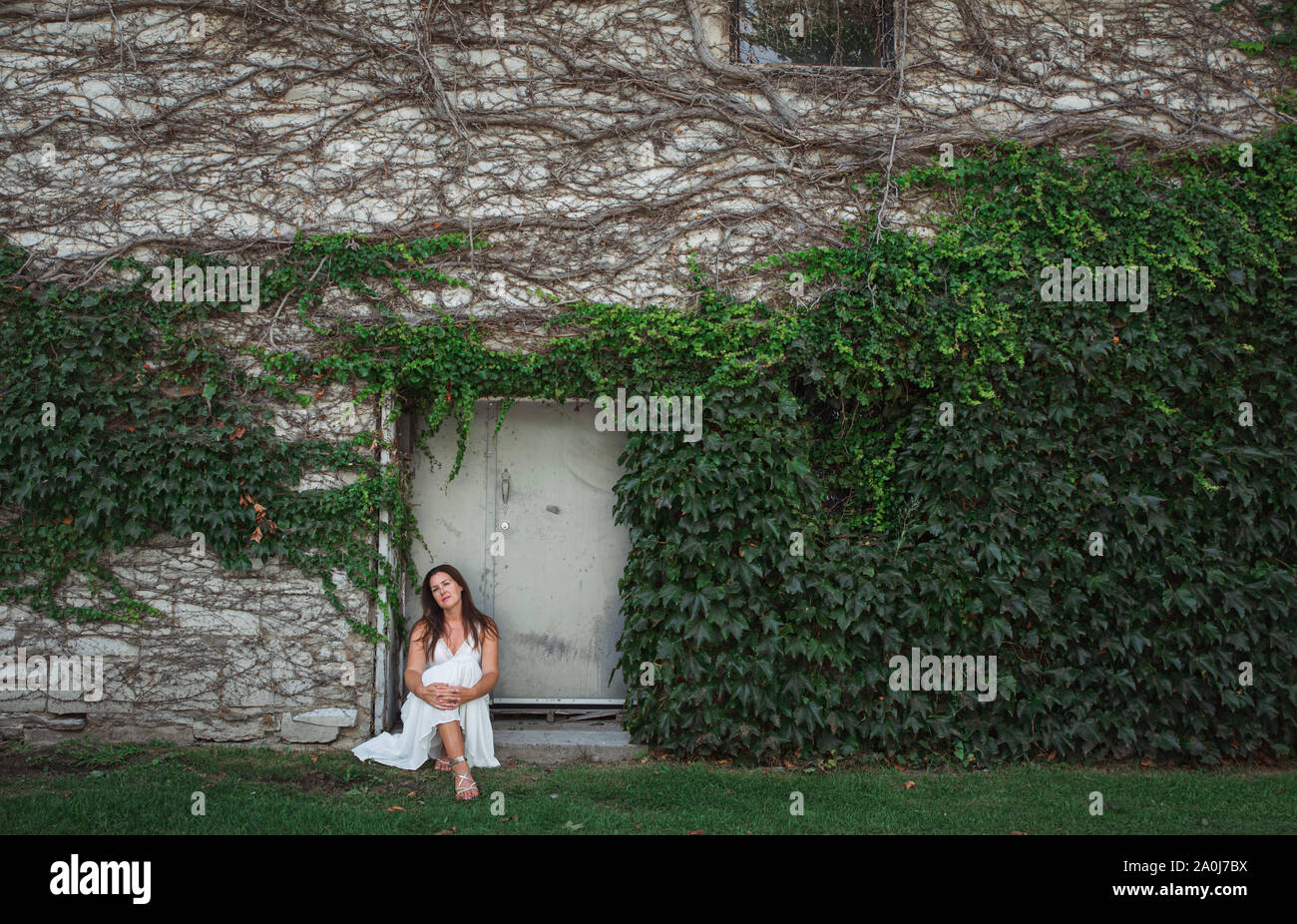 Femme en robe blanche assise dans l'embrasure d'un mur couvert de lierre. Banque D'Images