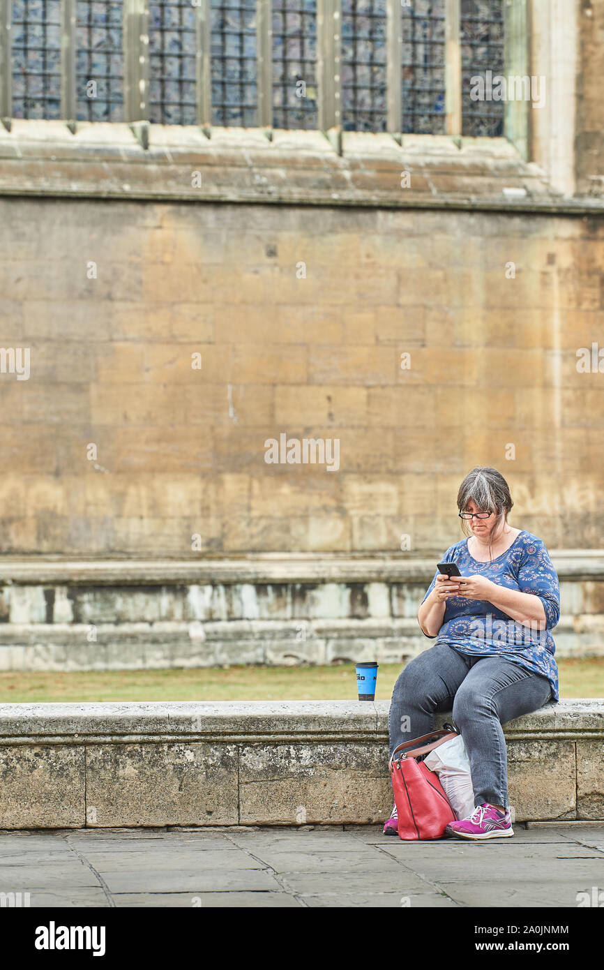Un puits construit femme regarde son téléphone alors qu'il était assis sur le muret à l'extérieur de la chapelle de King's College, Université de Cambridge, Angleterre. Banque D'Images
