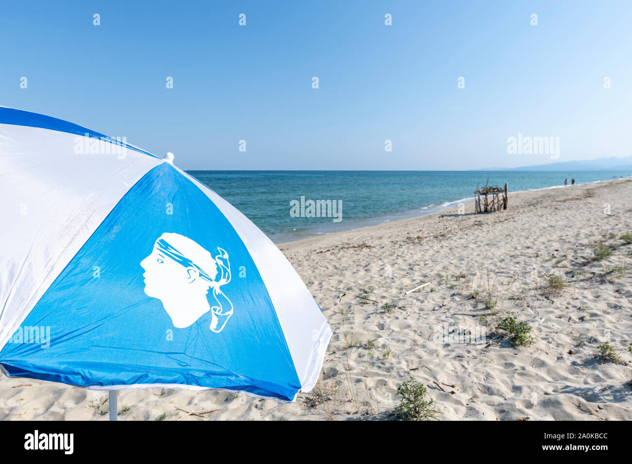 Drapeau corse sur un bleu et parapluie blanc contre une plage de sable et mer Méditerranée pendant l'été avec copie espace, Ghisonaccia, Corse, France Banque D'Images
