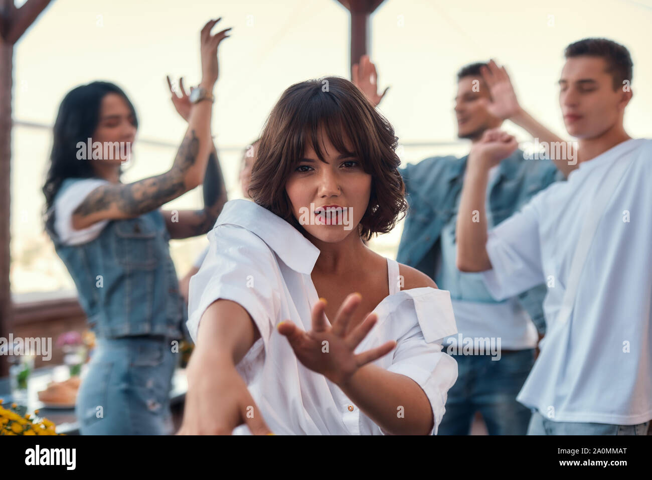 Se détendre. Jolie et jeune femme en chemise blanche est la danse et looking at camera en se tenant sur le toit-terrasse avec ses amis. Partie concept. L'amitié Banque D'Images