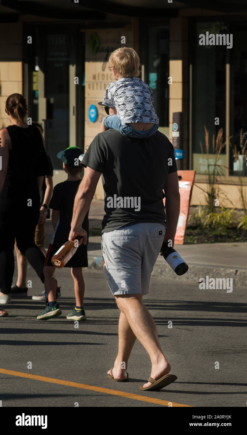Homme porte son enfant et deux bouteilles de vin sur l'avenue Maguire à Sillery, à la fin d'une foire de rue d'été au Québec, CA. Banque D'Images