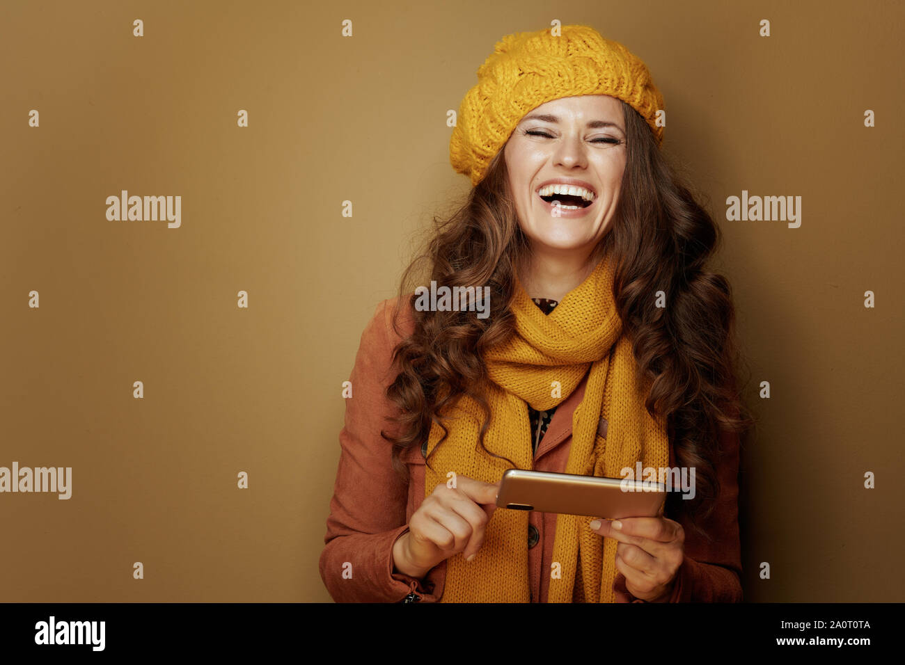 Portrait of smiling young woman in yellow beret et écharpe avec téléphone et SMS sur fond brun. Banque D'Images