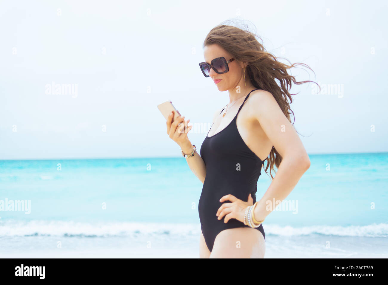 L'âge moyen élégant woman in sunglasses with phone texting sur une plage de sable blanc. Banque D'Images