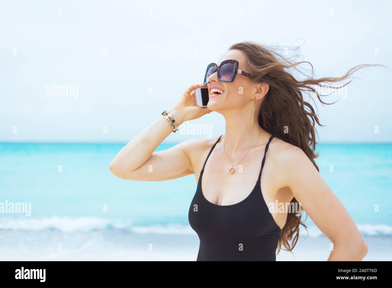 Moderne de 40 ans souriante woman in sunglasses sur une plage blanche parlant sur un téléphone cellulaire. Banque D'Images