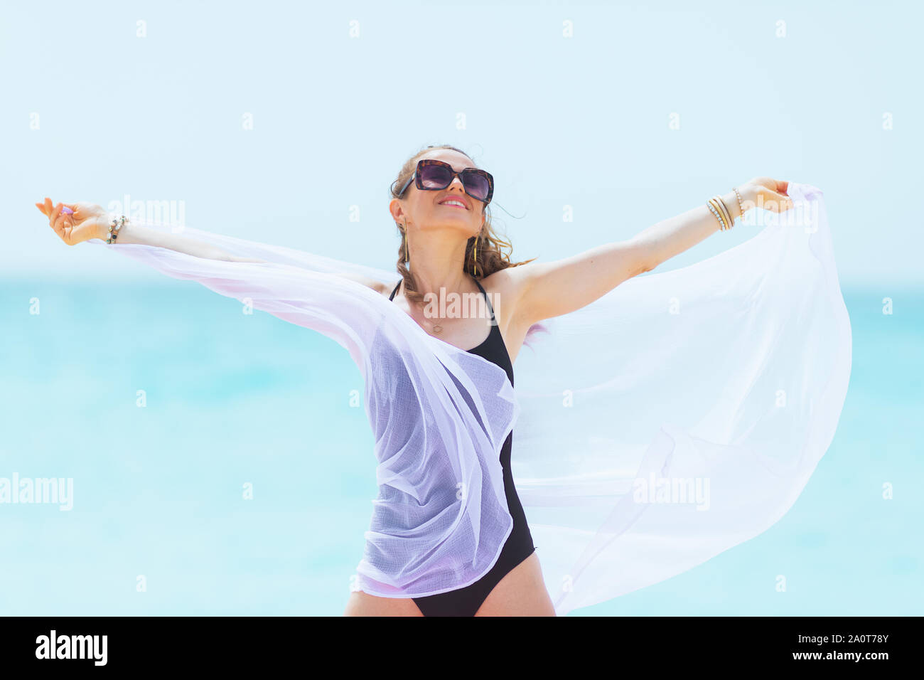 Cheerful young 40 ans femme aux longs cheveux bouclés en noir élégant maillot sur une plage blanche holding white paréos. Banque D'Images