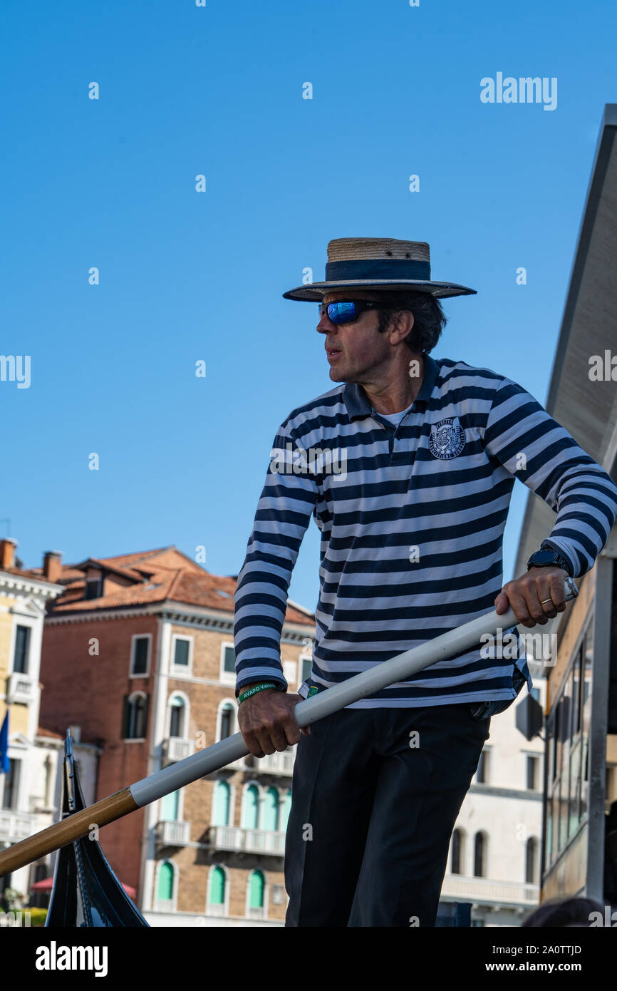 Un gondolier dans un chemise rayée et chapeau canotier de paille sur une gondole sur les canaux de Venise, Italie. Porter des lunettes de soleil miroir moderne. Banque D'Images
