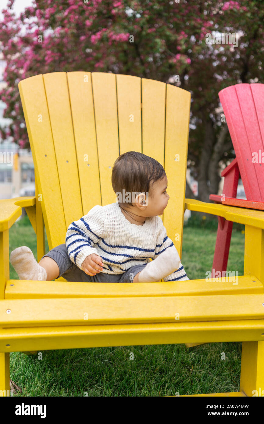 Bébé s'efforce de grimper sur l'accoudoir d'un fauteuil Muskoka debout sur une pelouse. Mignon enfant portant un chandail sur un ressort à pied. Banque D'Images