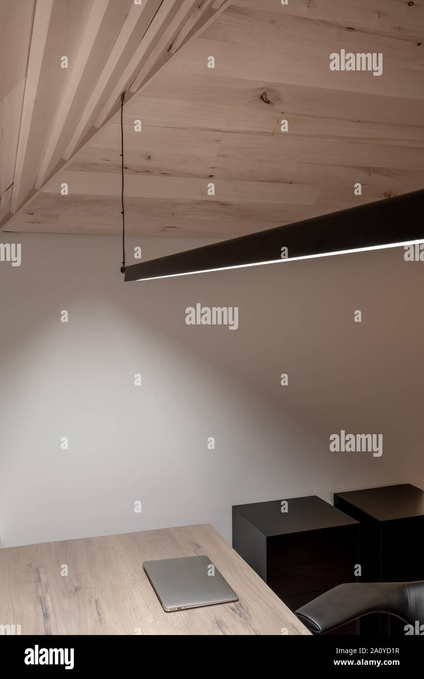 Intérieur contemporain avec une lumière en pente plafond en bois et de murs  blancs. Il y a une lampe lumineuse suspendue au-dessus de la table avec un  ordinateur portable, commod noir Photo