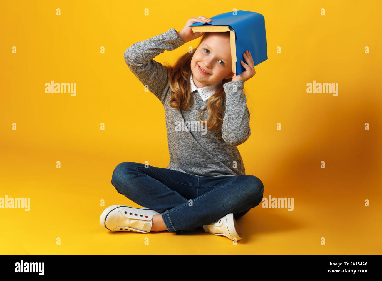 Une petite fille d'étudiant est assis sur le plancher avec un livre sur la tête. L'éducation et de l'école concept. Banque D'Images
