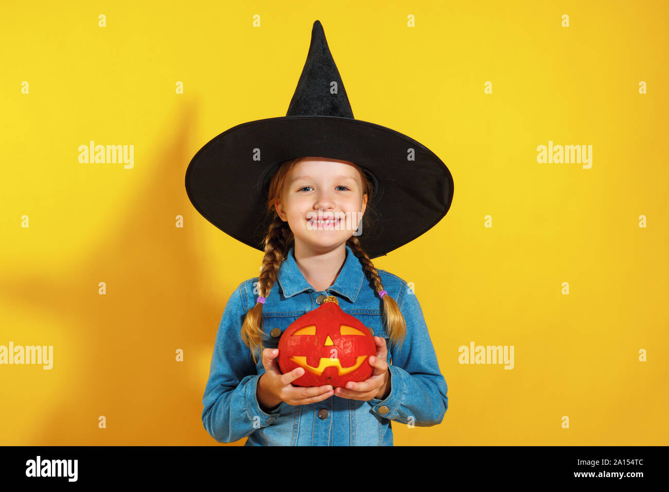 Portrait d'une petite fille dans un chapeau de sorcière. Un enfant est titulaire d'une citrouille Jack la lanterne sur un fond jaune. Célébration de l'Halloween. Banque D'Images