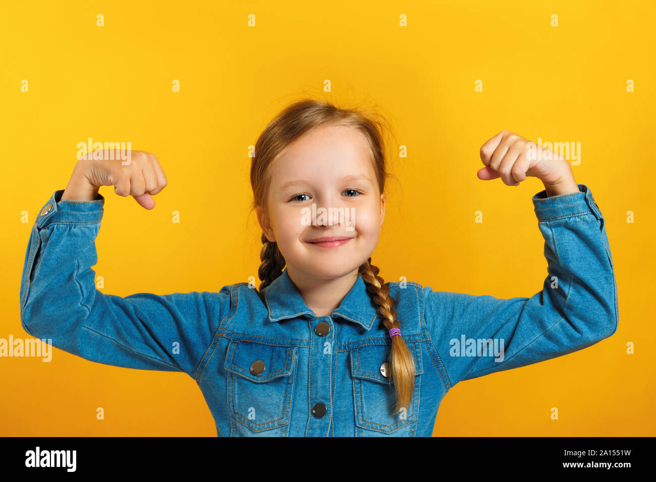 Petite fille dans une chemise en jean avec des nattes faites de poils sur un fond jaune. L'enfant montre des mains fortes. Concept de l'éducation, l'école, succès, powe Banque D'Images