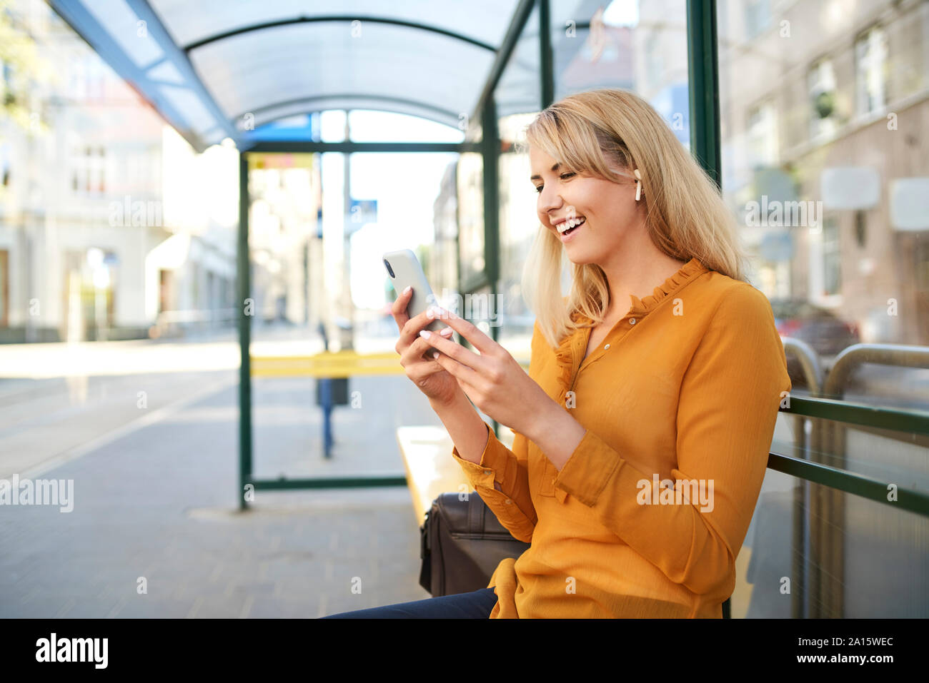 Happy young woman avec écouteurs sans fil à l'aide à l'arrêt de bus du smartphone Banque D'Images