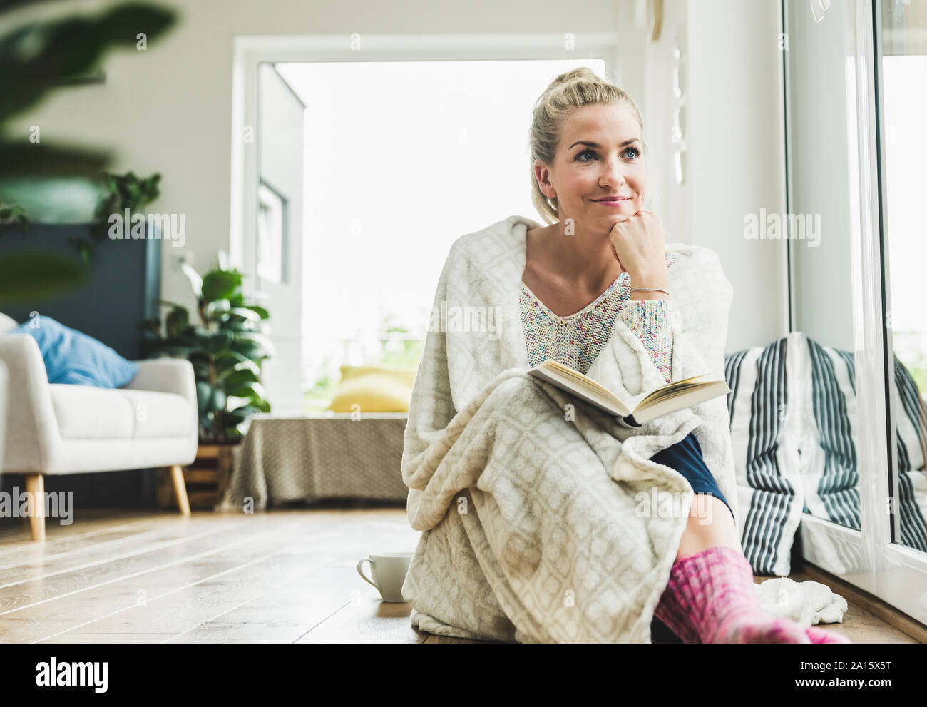 Femme enveloppée dans une couverture assis à la fenêtre à la lecture d'un livre Banque D'Images