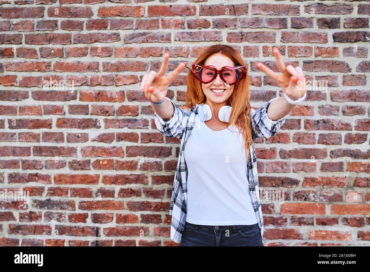 Cheerful young woman in heart-shaped glasses faisant la paix et gestes smiling at camera Banque D'Images