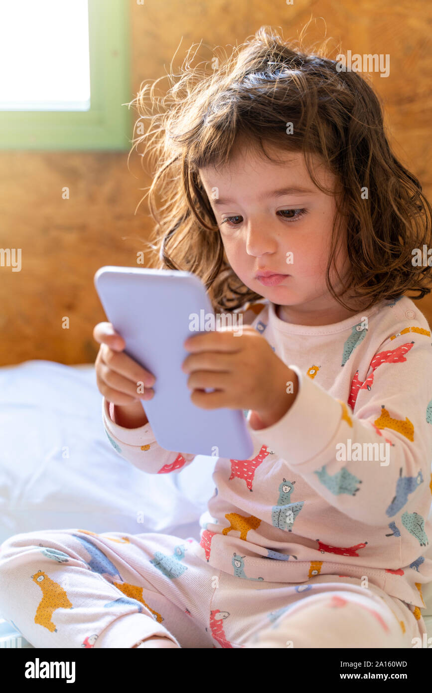 Cute little girl en pyjama dans le lit à l'aide de mobile phone Banque D'Images