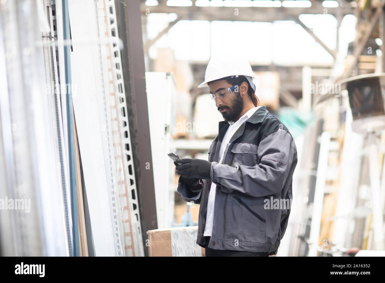 Jeune homme avec cell phone wearing hard hat travaillant dans un entrepôt Banque D'Images