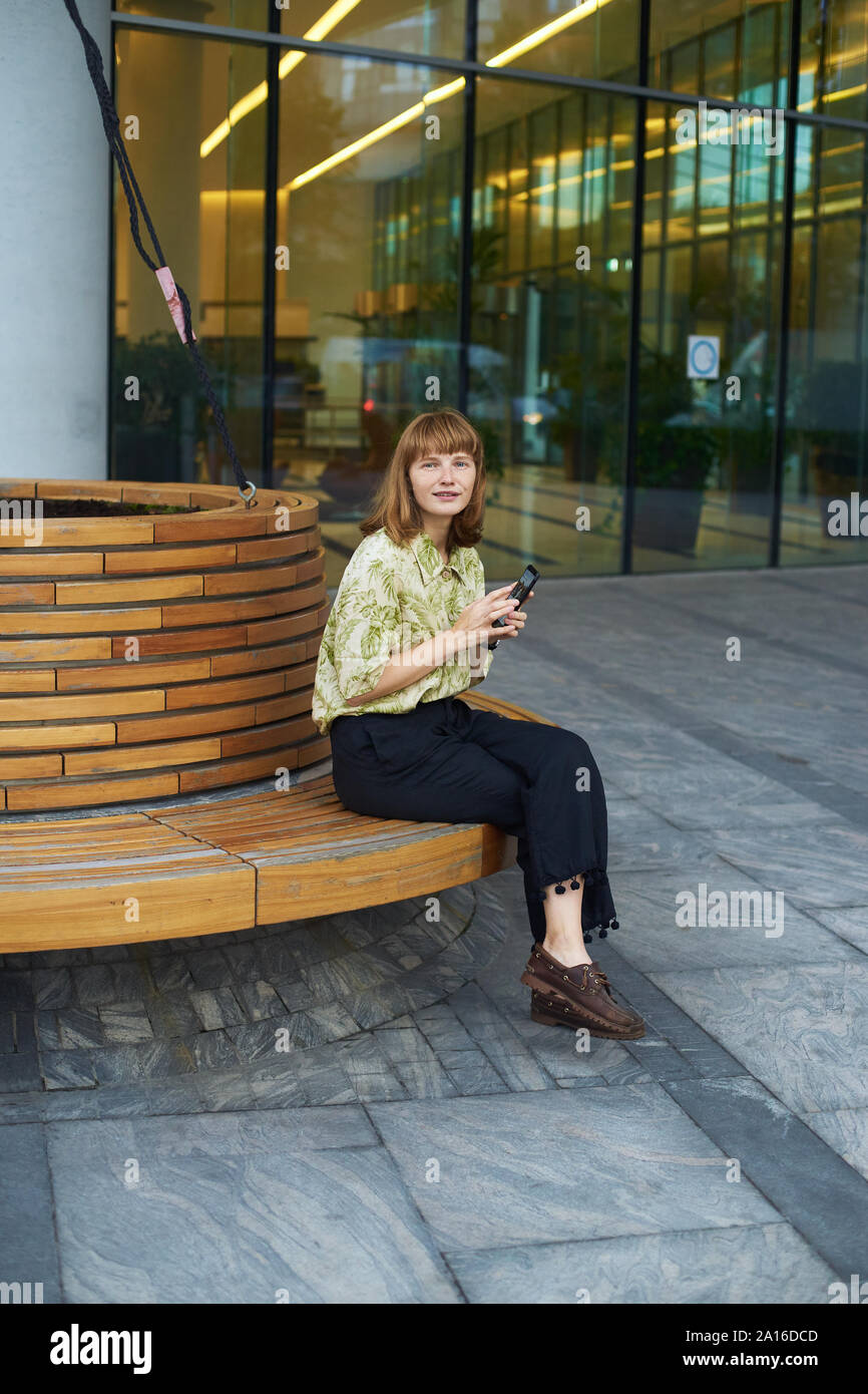 Jeune femme rousse de modèle avec rousseur assis au banc en bois près d'un centre d'affaires moderne et tenant son smartphone Banque D'Images
