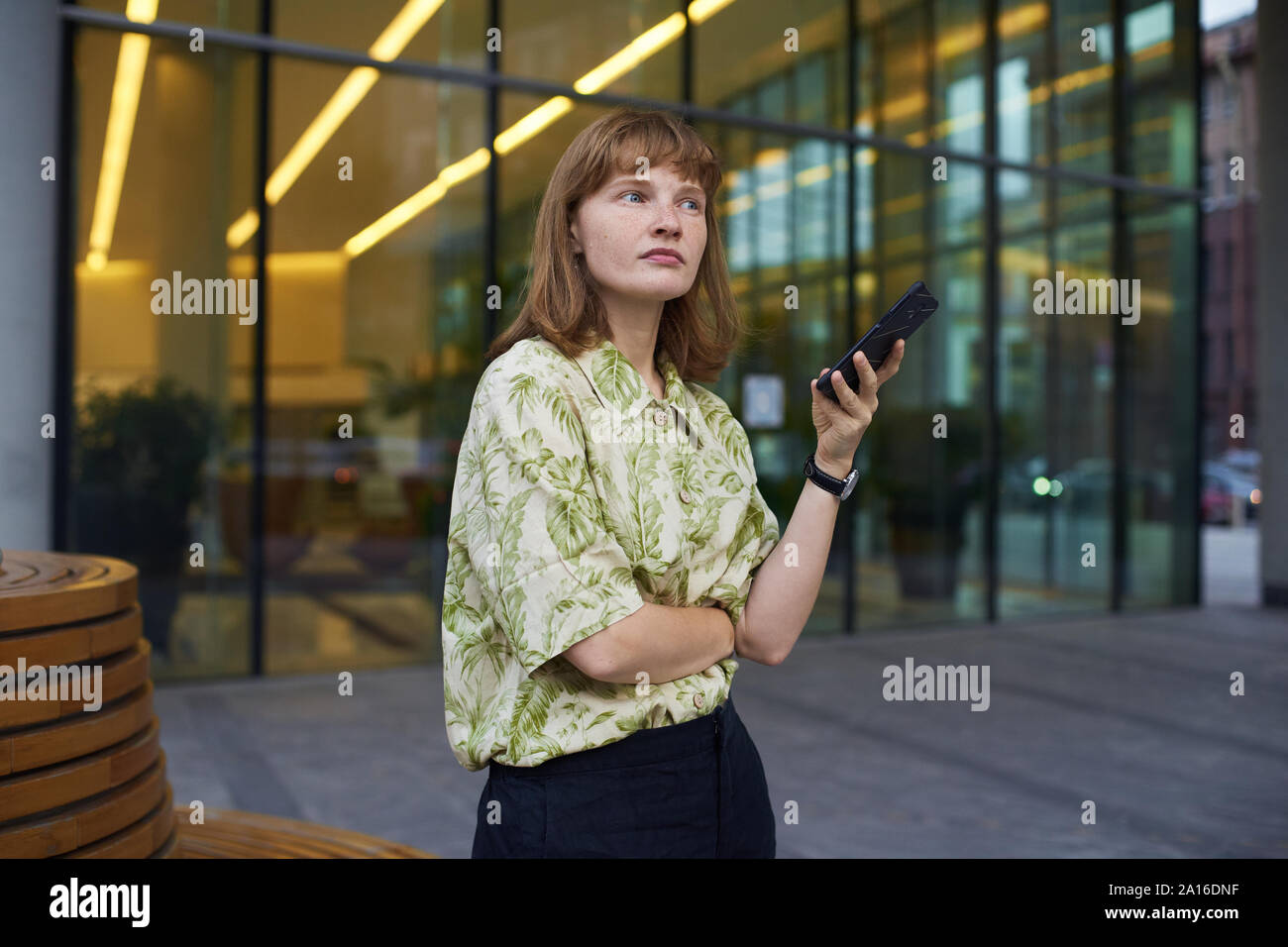Jeune femme avec des cheveux gingembre debout à la rue près d'un immeuble de bureaux tenant son téléphone et la pensée. La prise de décision Banque D'Images