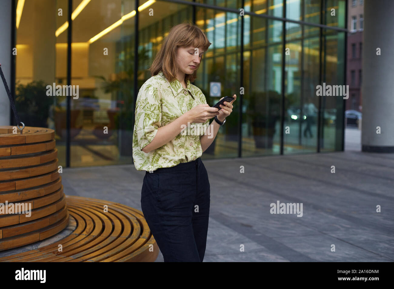 Jeune femme avec des cheveux gingembre debout à la rue près d'un immeuble de bureaux tenant son téléphone et la pensée Banque D'Images