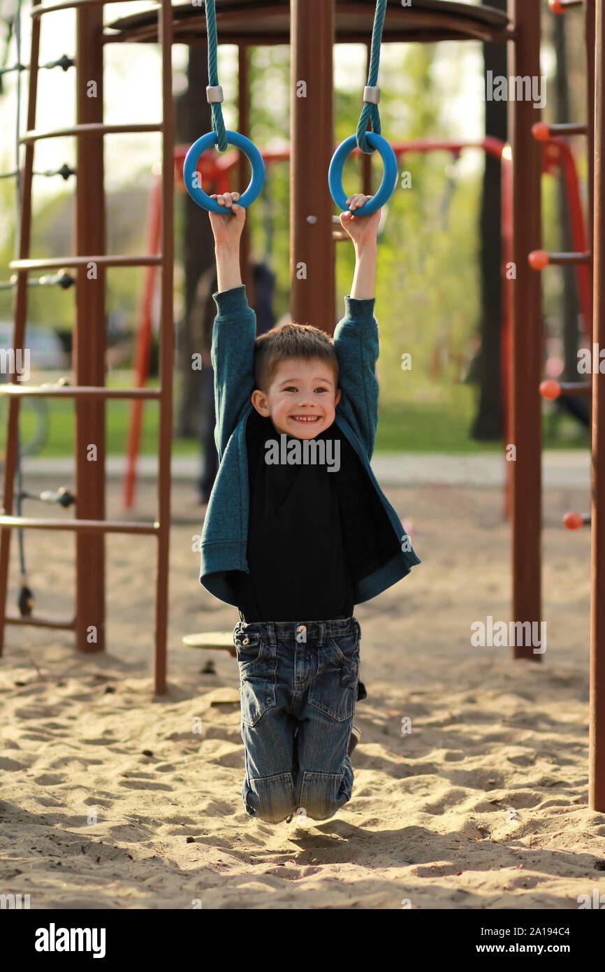 Happy smiling boy hanging on gymnastic rings sur l'aire de jeux extérieure. Portrait candide full size Banque D'Images