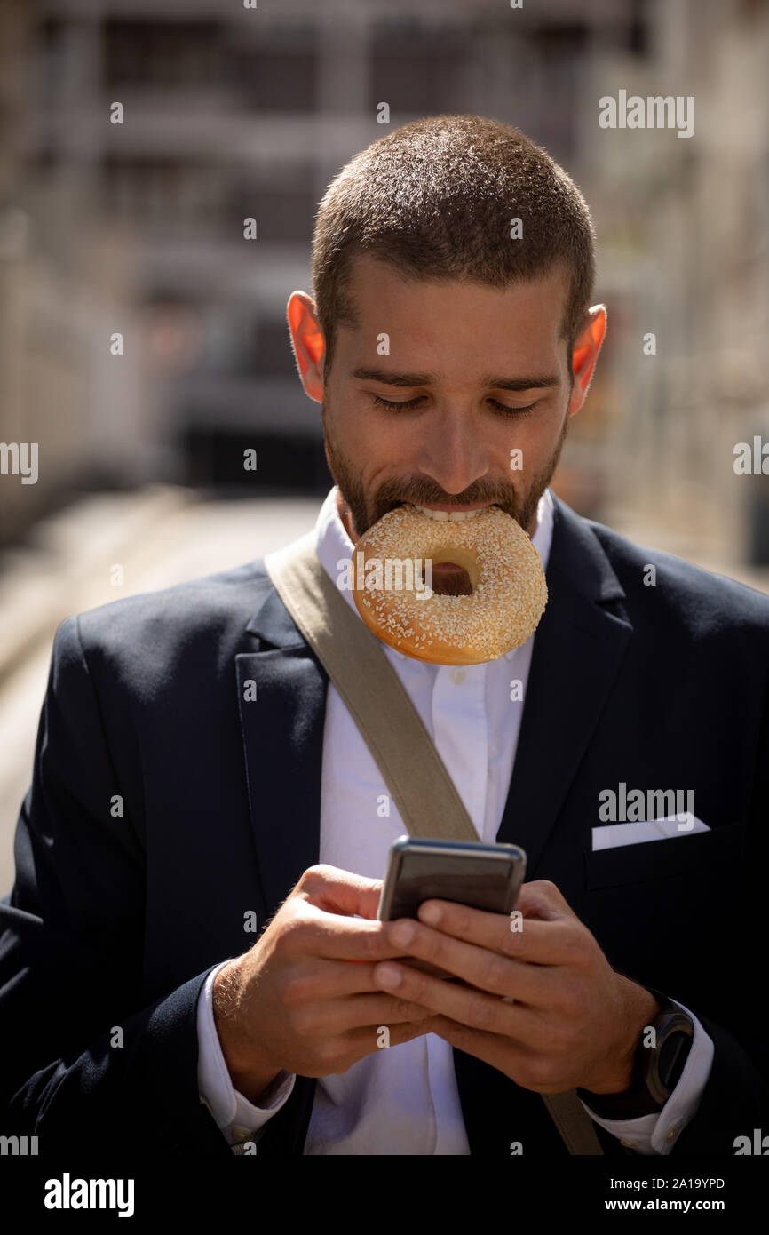 Jeune homme professionnel de manger un beigne et à l'aide du smartphone sur la rue Banque D'Images