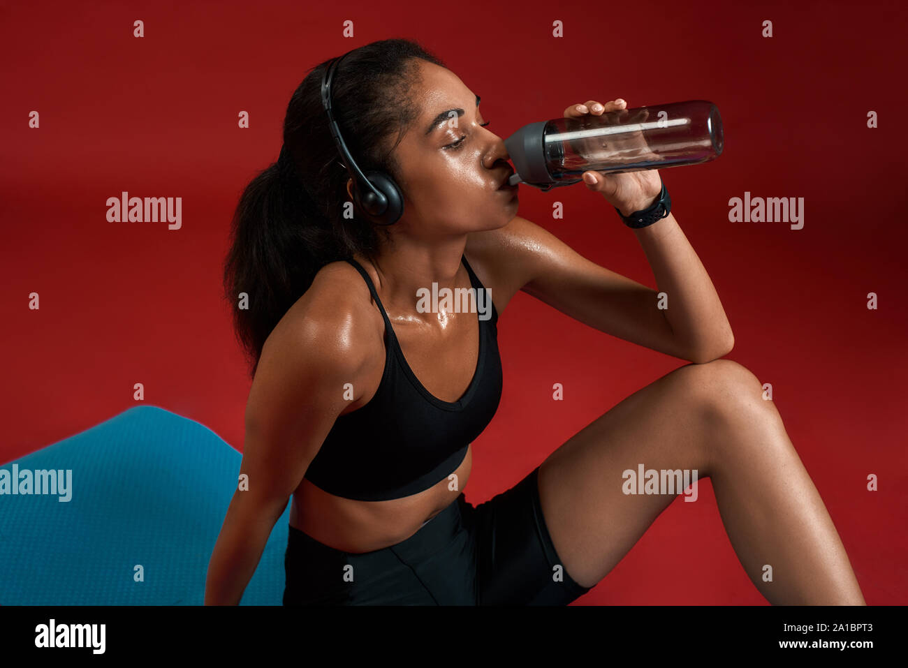 N'oubliez pas de boire suffisamment. Épuisé belle et jeune femme dans l'eau potable de sportswear tout en s'asseyant sur tapis d'exercice contre fond rouge. Concept sport Banque D'Images