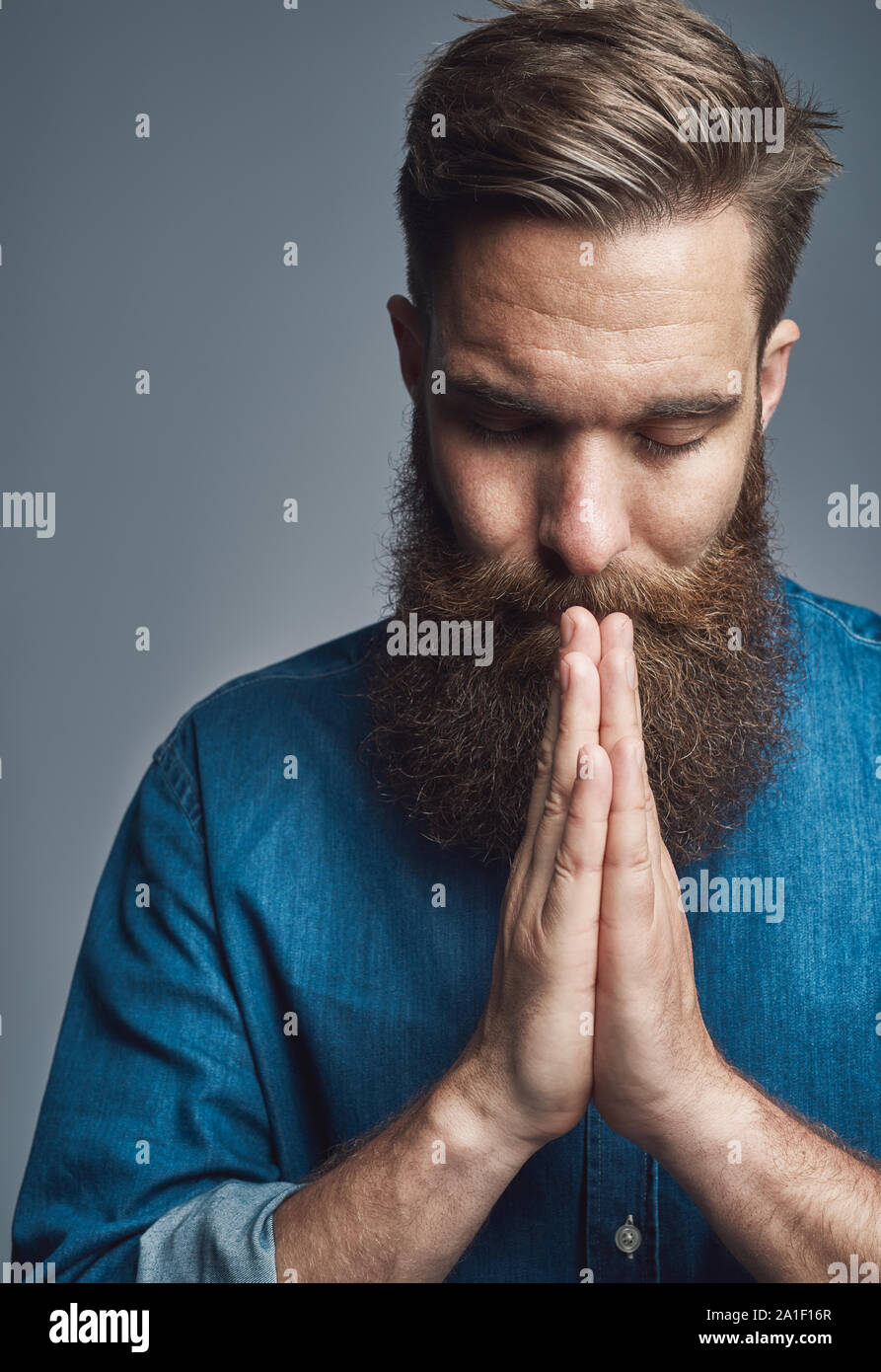 Jeune homme avec une longue barbe debout avec ses mains jointes en prière sur un arrière-plan gris Banque D'Images