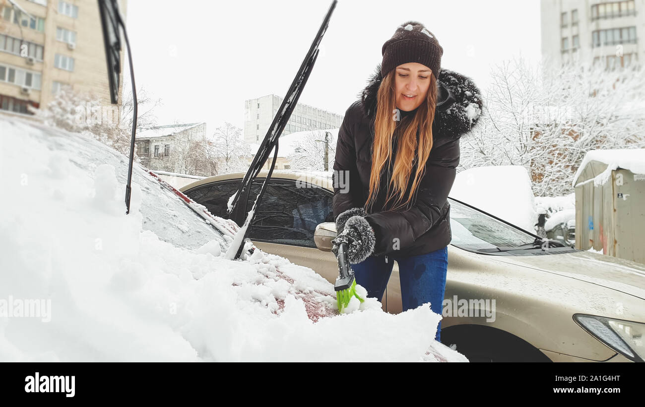 La photo en gros plan de jeunes et belles smiling blonde femme essayant de nettoyer les couverts de neige par automatique rouge brosse après une tempête de neige. Gratter le pare-brise et Banque D'Images