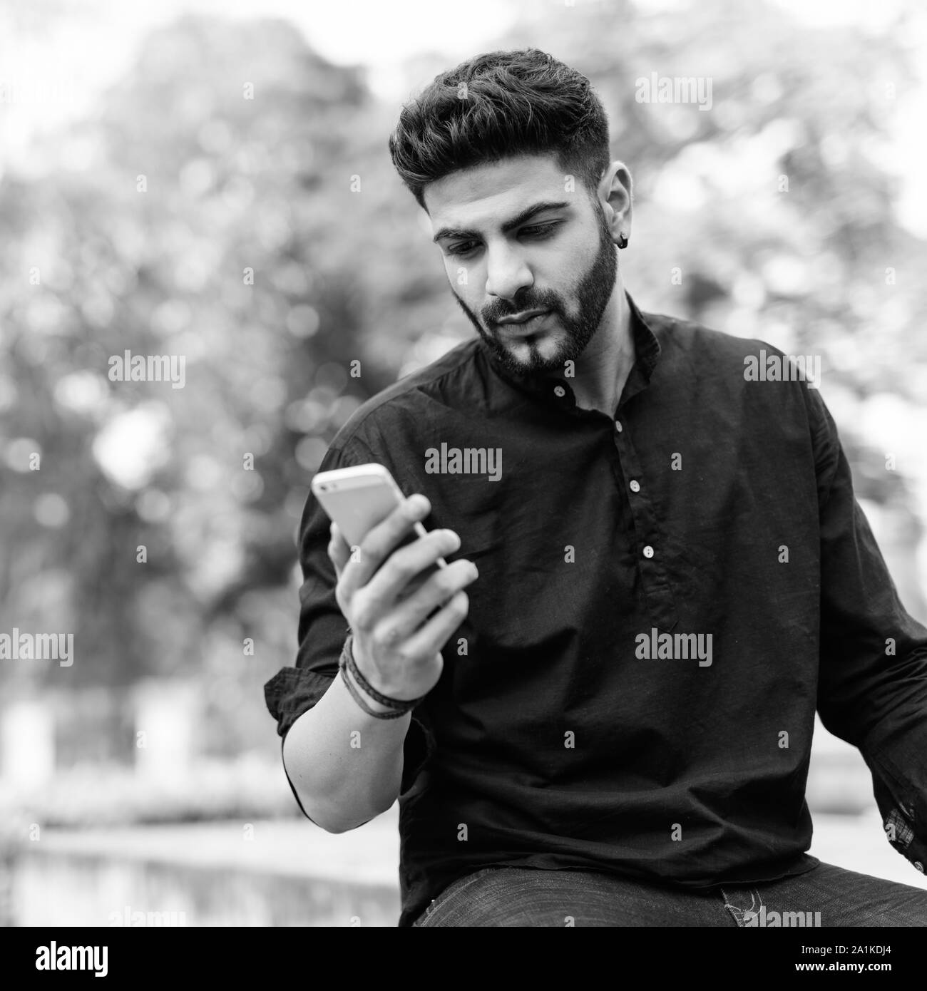 Beau jeune homme indien à l'aide de téléphone mobile tout en restant assis sur un sol en béton au parc Banque D'Images