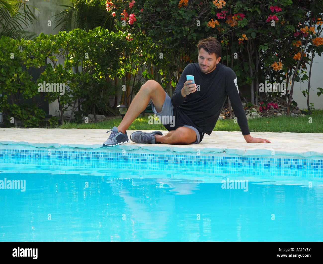 Un homme dans les vêtements de sport, assis près de la piscine dans l'hôtel, utilise un téléphone cellulaire. Banque D'Images