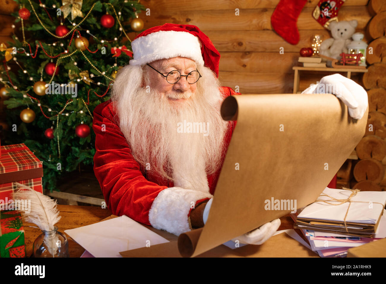 Costume Père Noël en le déroulant grand papier avec voeux de Noël assis par table en bois à la maison Banque D'Images