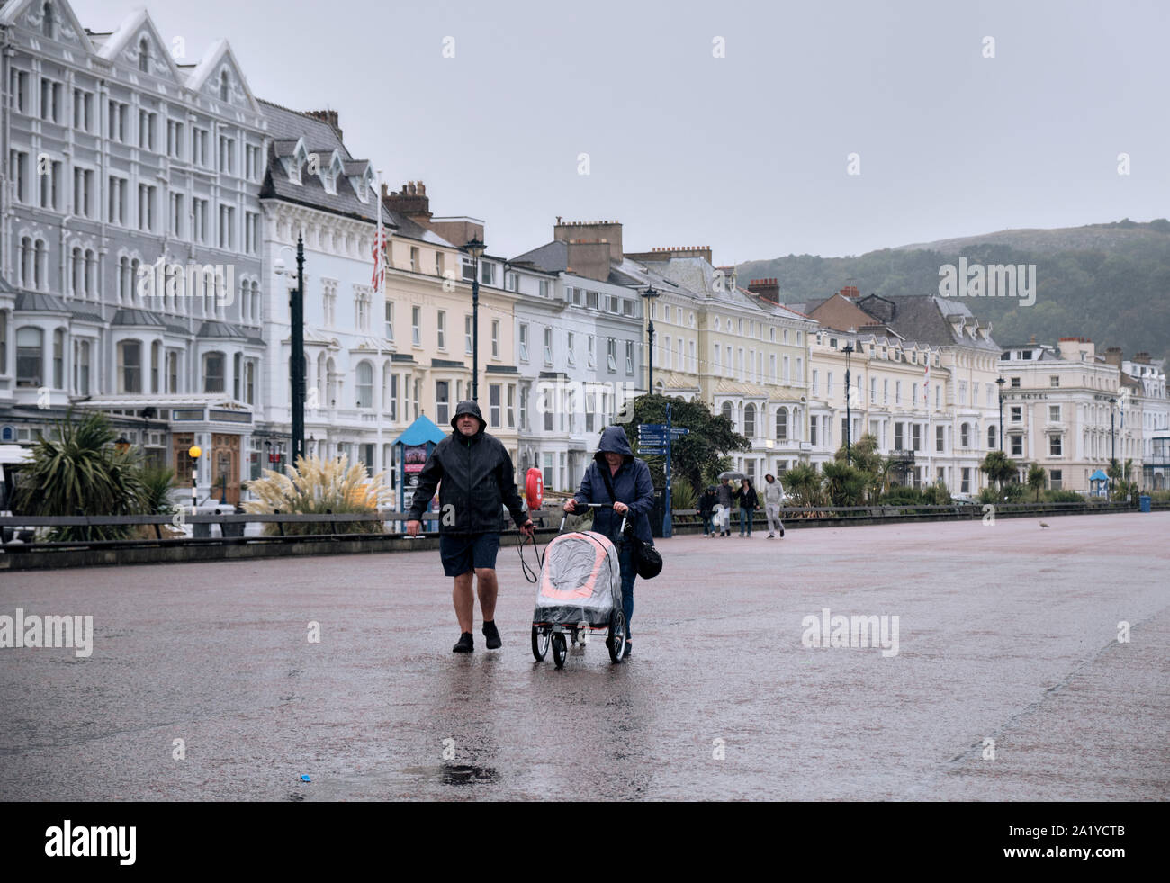 En quelques vêtements de pluie poussant un landau, une marche sous la pluie sur l'esplanade à Llandudno Pays de Galles sur un jour d'automne gris Banque D'Images