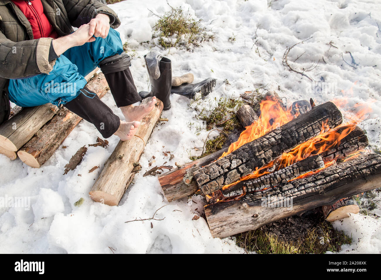 Un homme se réchauffe les mains et les pieds près du feu dans la forêt de pins couverts de neige en hiver Banque D'Images