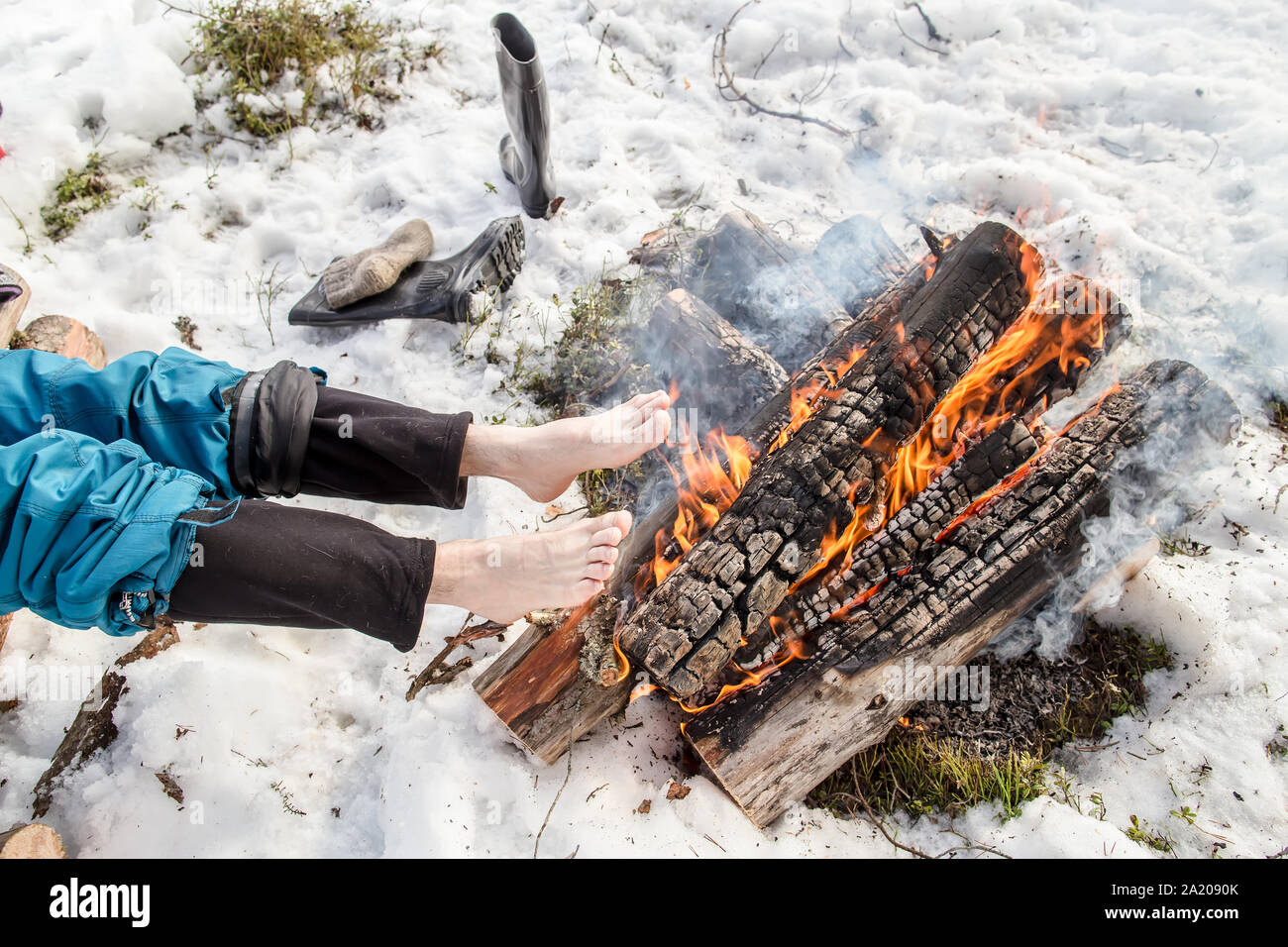 Un homme se réchauffe ses pieds nus près de l'incendie dans la forêt de pins couverts de neige en hiver Banque D'Images