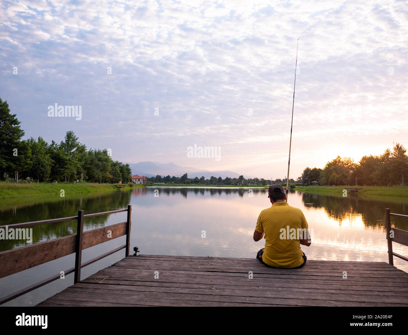 Il y a une porte jaune homme tenant un hameçon assis sur le bord d'un quai près d'un petit lac au milieu forrest pendant le coucher du soleil Banque D'Images