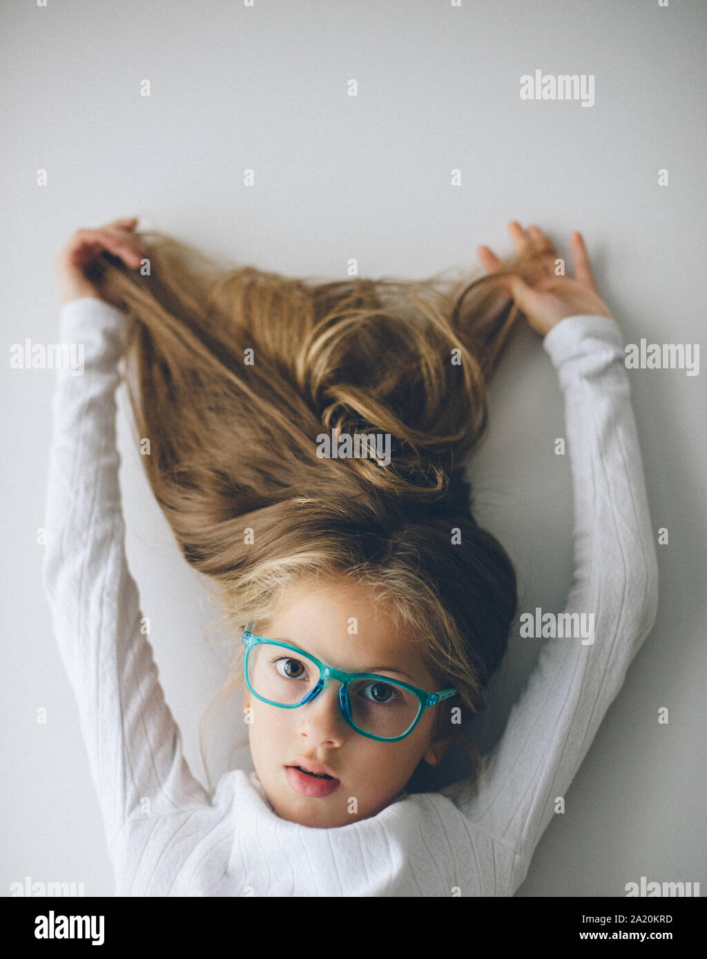 Belle petite caucasian girl in blue glasses holding ses longs cheveux blonds jusqu' Banque D'Images