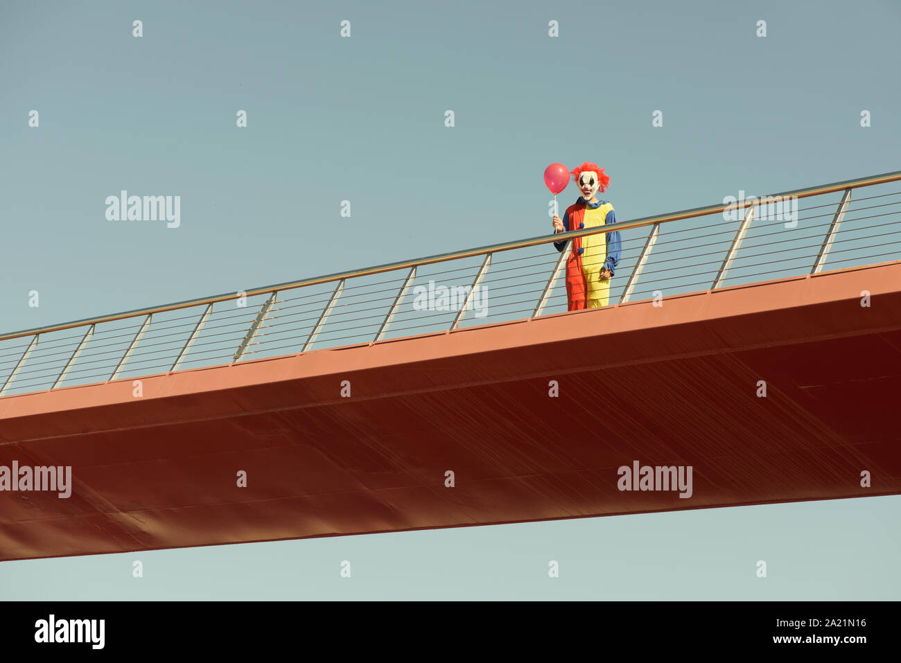 Un clown effrayant colorés portant un jaune, rouge et bleu costume, tenant un ballon rouge dans sa main, debout dans un pont en plein air Banque D'Images