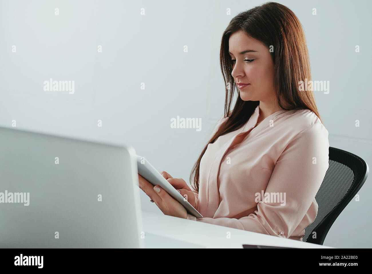 Beau young businesswoman sitting in front of laptop using digital tablet in office Banque D'Images