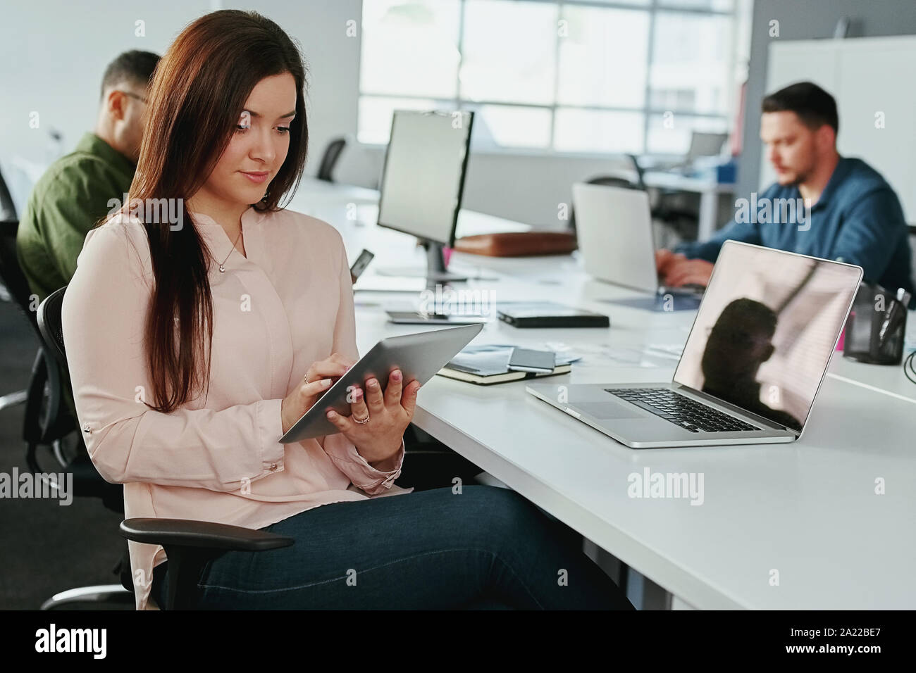 Un séduisant jeune smiling businesswoman using digital tablet avec des collègues travaillant au bureau en arrière-plan à l'office de tourisme - Office de démarrage Banque D'Images