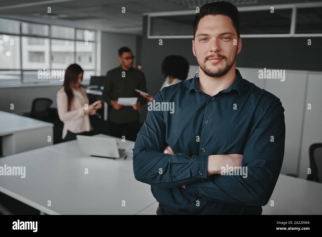 Beau jeune homme professionnel de bureau avec bras croisés à la caméra et à d'autres travailleurs ayant conversation en arrière-plan Banque D'Images