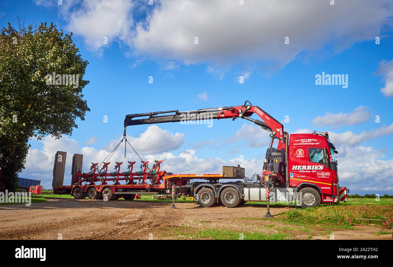 Un Transport Ltd rouge Herbies Volvo FH13.520 camion grue camion avec remorque surbaissée de machines agricoles de déchargement avec ses béquilles et flèche déployée Banque D'Images