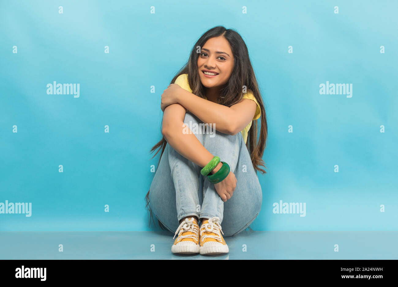 Smiling teenage girl en jeans et t-shirt assise sur le sol tenant ses genoux avec les mains Banque D'Images