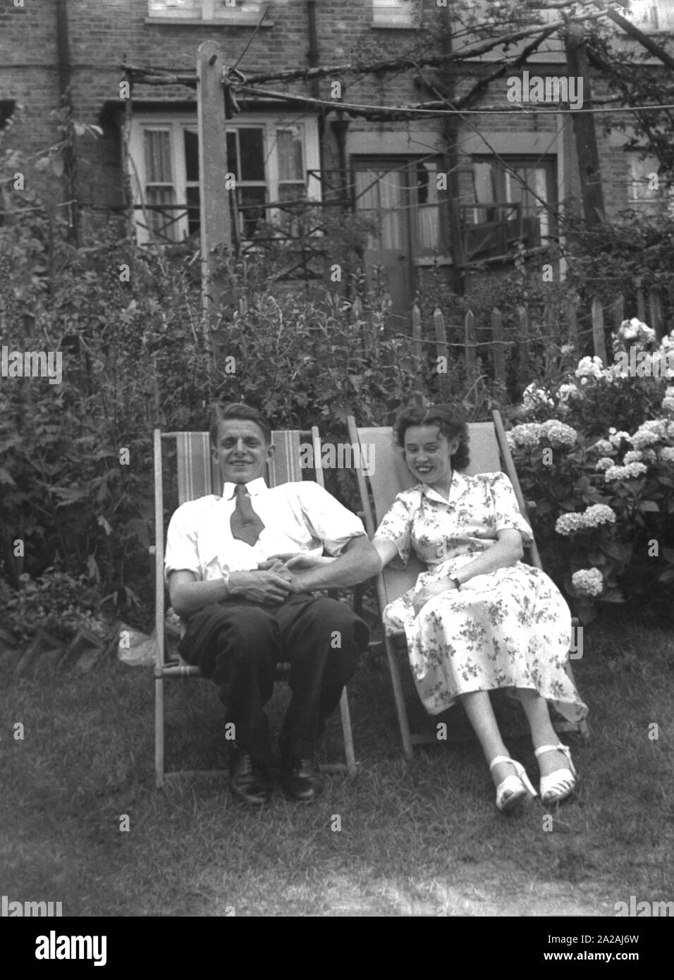 Années 1950, historique, un couple assis ensemble sur des transats dans un jardin arrière tenant la main, Angleterre, Royaume-Uni. Banque D'Images