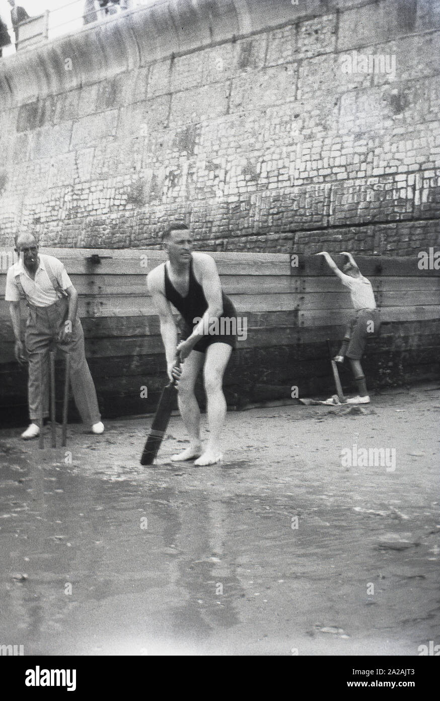 Années 1930, historique, hommes jouant au cricket sur une plage près d'un brise-mer et d'un mur géant de la plage, batteur dans son maillot de bain sans manches, Angleterre, Royaume-Uni. Banque D'Images