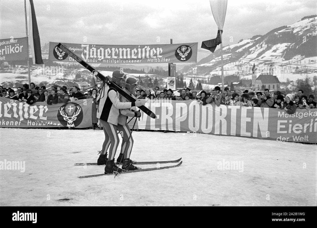 Deux coureurs se font passer pour la photographie des spectateurs sur une paire de skis après ils arrivent à la ligne d'arrivée. Il y accrocher de grandes bannières publicitaires des sponsors de la course dans l'aire d'arrivée, dans ce cas Jaegermeister et Verpoorten. La course du Hahnenkamm a eu lieu sur le Hahnenkamm de Kitzbühel depuis 1931. Banque D'Images