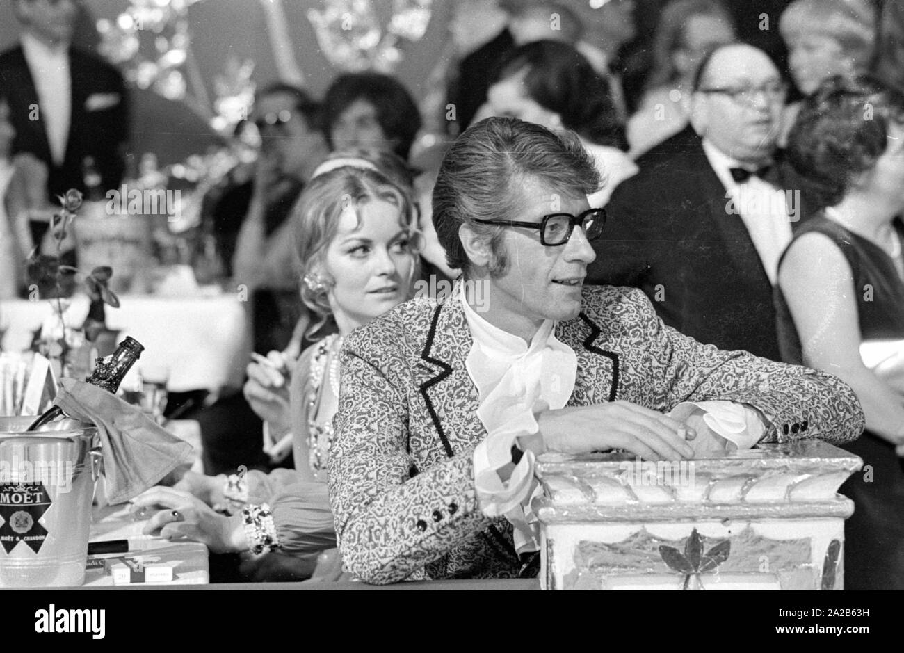 Rudi Carrell assis à une table remplie de champagne et verres sur le film Munich, balle dans le Bayerischer Hof. Carrell porte une veste à motifs et une chemise froissée, il s'appuie sur un pilier. Banque D'Images