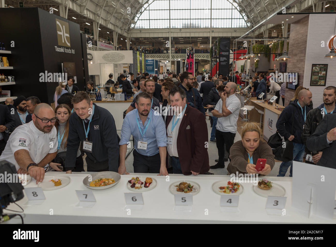 Olympia, Londres, Royaume-Uni. 2 octobre 2019. Une compétition culinaire ou une exposition gastronomique organisée dans un grand lieu couvert avec un haut plafond voûté. Huit boîtes plaquées sont affichées sur un compteur d'évaluation, chacune étiquetée numériquement. Les juges ou les participants se tiennent derrière le comptoir pour évaluer la présentation. Une personne photographie la vaisselle avec un smartphone. L’arrière-plan présente des stands d’exposition animés et des participants, soulignant la popularité de l’événement et l’engagement du public. Penelope Barritt/Alamy Live News Banque D'Images