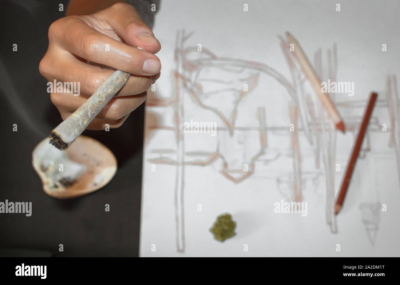 Fumer un joint de marijuana pendant une pause après le dessin. Close up of hands et le joint, l'effet de flou. Arrière-plan avec des crayons et des croquis, copyspace. Banque D'Images